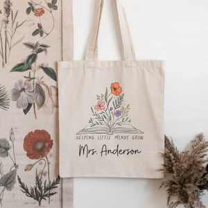 May include: A beige canvas tote bag featuring a black line drawing of an open book with colorful flowers blooming from the pages. Below the illustration, the text reads "HELPING LITTLE MINDS GROW" in capital letters and "Mrs. Anderson" in script.