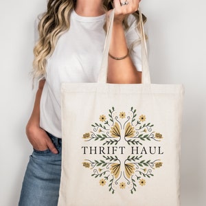 Pu&ograve; includere: Una borsa tote in tela bianca con un design floreale e il testo "Thrift Haul".