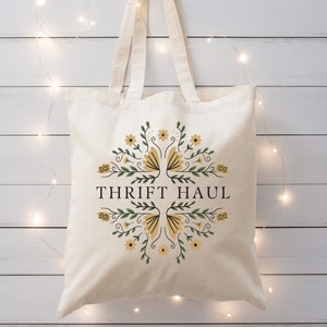 Pu&ograve; includere: Una borsa tote in tela bianca con un design floreale in tonalit&agrave; gialle e verdi. Il testo "THRIFT HAUL" &egrave; stampato in nero sulla borsa.