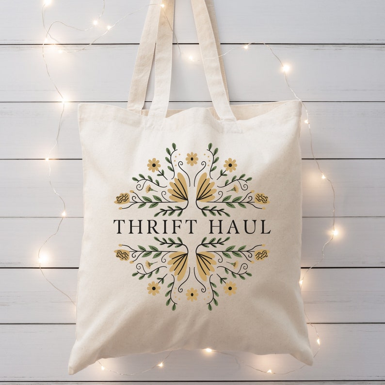 Pu&ograve; includere: Una borsa tote in tela bianca con un design floreale in tonalit&agrave; gialle e verdi. Il testo "THRIFT HAUL" &egrave; stampato in nero sulla borsa.