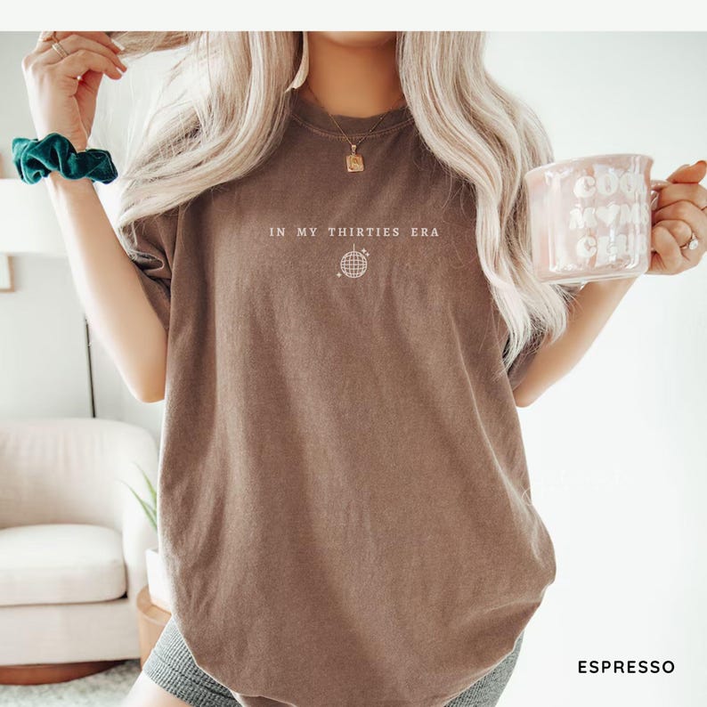 以下が含まれることがあります： エスプレッソカラーの半袖Tシャツ。胸の中央に白い文字で「IN MY THIRTIES ERA」とプリントされ、その下に小さな白いミラーボールのグラフィックが描かれています。クルーネックでゆったりとしたフィット感のブラウンの生地です。