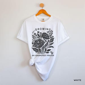 May include: A white short-sleeve t-shirt featuring a dark gray graphic print of a floral arrangement with the text "GROWING" above and "AN ANSWERED PRAYER" below, accompanied by two small baby footprints near the bottom of the floral design.