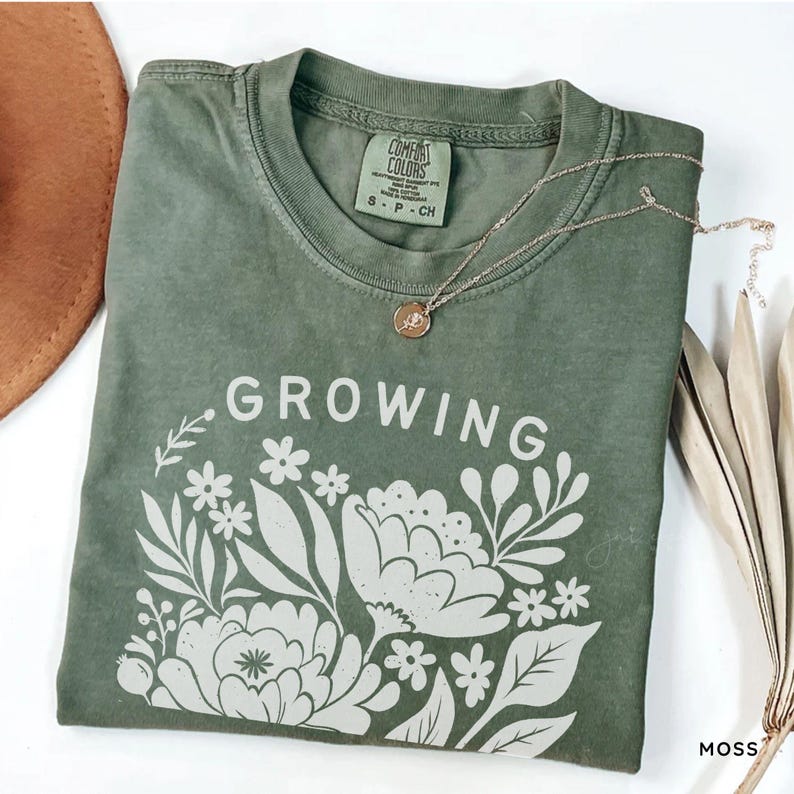 May include: An olive green short-sleeve t-shirt with a crew neckline. The shirt features a white graphic print of flowers and leaves with the text "GROWING" above the floral design. A Comfort Colors brand label is visible at the inner neckline.