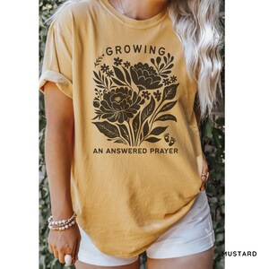 May include: A mustard yellow short-sleeve t-shirt featuring a dark brown graphic of a floral bouquet. The text "GROWING" is printed above the flowers, and "AN ANSWERED PRAYER" is printed below, accompanied by a small icon of baby footprints.
