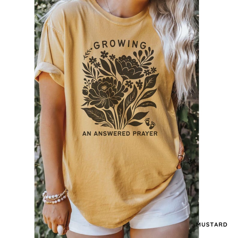 May include: A mustard yellow short-sleeve t-shirt featuring a dark brown graphic of a floral bouquet. The text "GROWING" is printed above the flowers, and "AN ANSWERED PRAYER" is printed below, accompanied by a small icon of baby footprints.