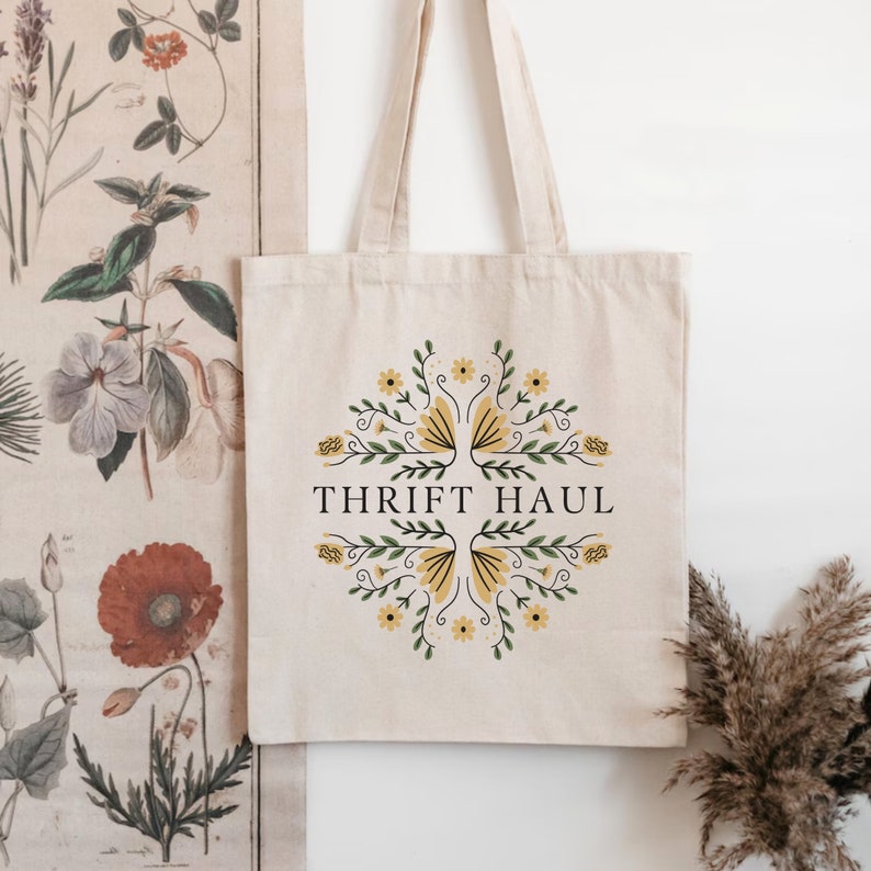 Pu&ograve; includere: Una borsa tote beige con un design floreale e il testo "Thrift Haul" stampato sul davanti.
