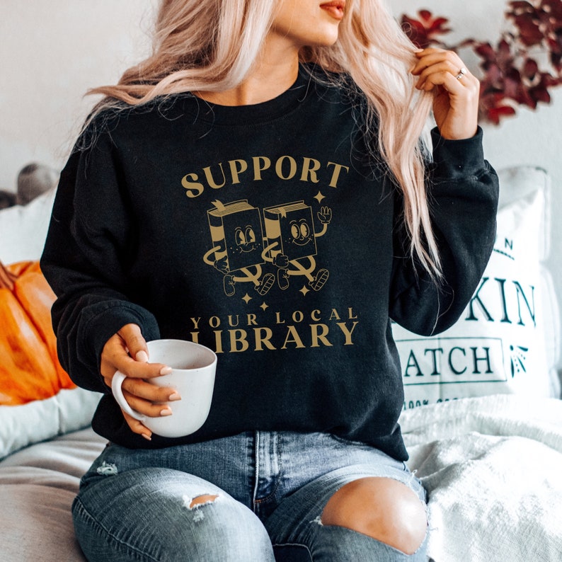 Puede incluir: Una sudadera negra con el texto "SUPPORT YOUR LOCAL LIBRARY" en letras doradas. Dos libros de dibujos animados se toman de la mano y sonr&iacute;en.