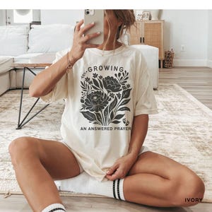 May include: An ivory short-sleeve t-shirt featuring a black graphic print of a floral bouquet with the text "GROWING" at the top and "AN ANSWERED PRAYER" at the bottom, accompanied by two small baby footprint icons near the base of the flowers.