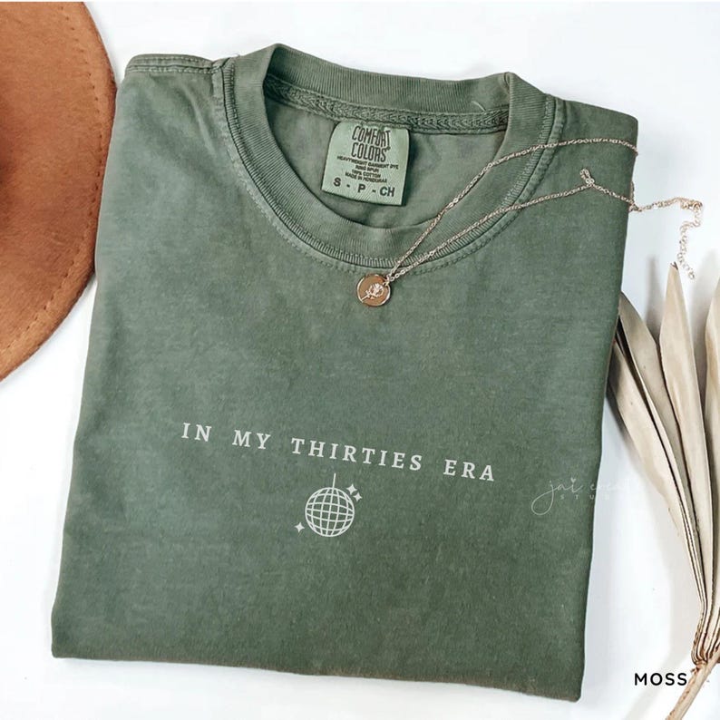 以下が含まれることがあります： モスグリーンの半袖Tシャツで、クルーネックとComfort Colorsのブランドラベルが付いています。フロントには白い文字で「IN MY THIRTIES ERA」と書かれ、その下にミラーボールのグラフィックが描かれています。小さな丸いペンダントが付いた細いゴールドトーンのチェーンネックレスが生地の上に置かれています。
