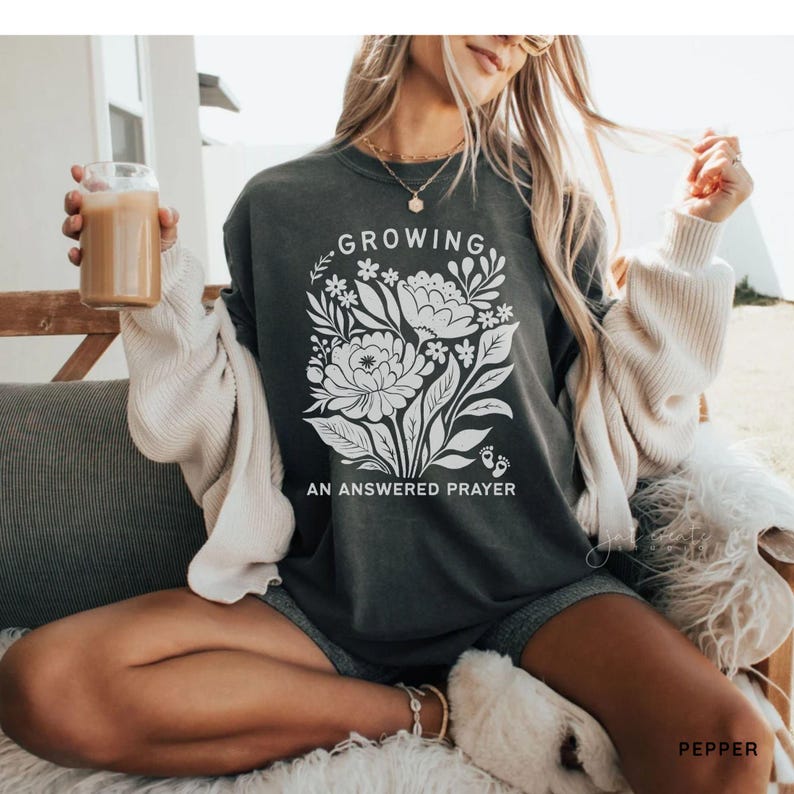 May include: A dark charcoal gray crewneck sweatshirt featuring a white graphic of a floral bouquet with the text "GROWING AN ANSWERED PRAYER" and two small baby footprints. The sweatshirt is worn with a cream-colored cardigan over the shoulders.