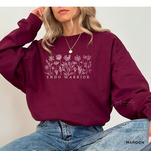 Pu&ograve; includere: Una felpa color bordeaux con un disegno floreale bianco e il testo "ENDO WARRIOR".