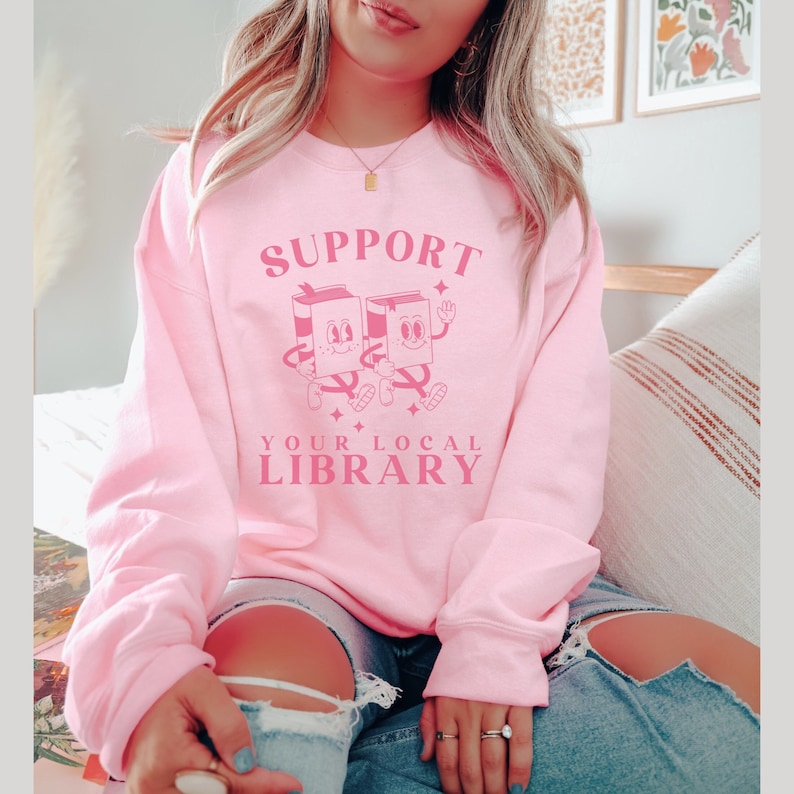 Puede incluir: Una sudadera rosa con el texto "SUPPORT YOUR LOCAL LIBRARY" en letras rosas. Dos libros de dibujos animados con caras se toman de la mano y caminan.