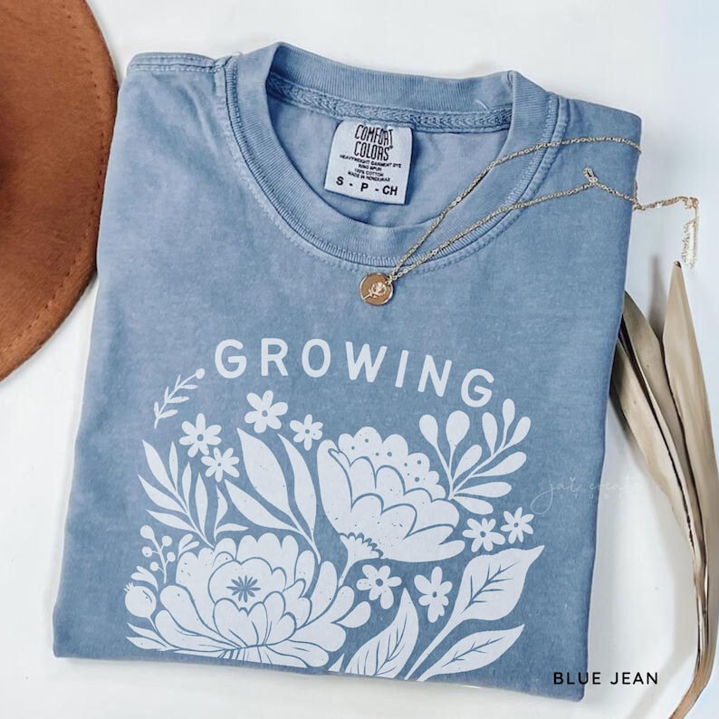 May include: A blue jean colored t-shirt with a white graphic print featuring the word GROWING above a floral arrangement. The shirt has a crew neckline and a Comfort Colors brand label on the inside collar. A gold chain necklace rests on the fabric.