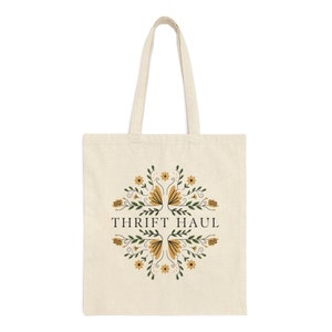 Pu&ograve; includere: Una borsa tote beige con un design floreale e il testo "Thrift Haul" in nero.