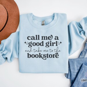 Może przedstawiać: Jasnoniebieska bluza z okrągłym dekoltem z ciemnoszarym napisem "call me a good girl and take me to the bookstore" oraz małymi akcentami w kształcie gwiazdek. Odzież jest pokazana obok brązowego filcowego kapelusza i niebieskich dżins&oacute;w.