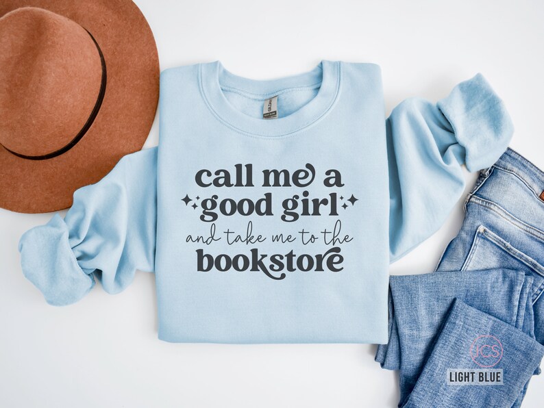 Może przedstawiać: Jasnoniebieska bluza z okrągłym dekoltem z ciemnoszarym napisem "call me a good girl and take me to the bookstore" oraz małymi akcentami w kształcie gwiazdek. Odzież jest pokazana obok brązowego filcowego kapelusza i niebieskich dżins&oacute;w.