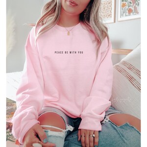 Pu&ograve; includere: Una felpa rosa con la scritta "Peace be with you" stampata sul davanti.
