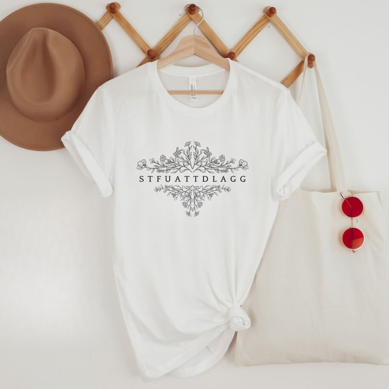 May include: White t-shirt with a floral design and the text "STFUATTDLAGG" printed on the front.