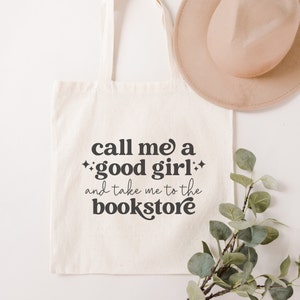 May include: A cream-colored canvas tote bag featuring the black printed text "call me a good girl and take me to the bookstore" with small star icons. The bag has two shoulder straps and is displayed alongside a tan hat and green foliage.