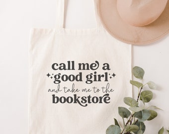 May include: A cream-colored canvas tote bag featuring the black printed text "call me a good girl and take me to the bookstore" with small star icons. The bag has two shoulder straps and is displayed alongside a tan hat and green foliage.