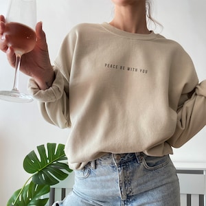 Pu&ograve; includere: Una felpa beige con la scritta "Peace be with you" stampata sul davanti. La felpa &egrave; indossata da una persona che indossa jeans blu.