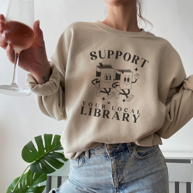 Puede incluir: Una sudadera beige con el texto "SUPPORT YOUR LOCAL LIBRARY" y un gr&aacute;fico de dos libros de dibujos animados con caras.