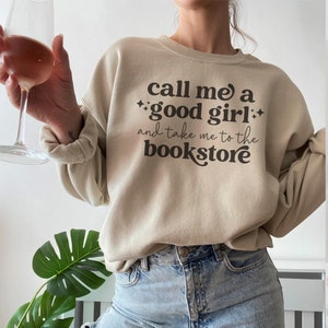 Może przedstawiać: Beżowa bluza z okrągłym dekoltem i ciemnobrązowym napisem call me a good girl and take me to the bookstore, ozdobiona małymi czteroramiennymi gwiazdkami. Odzież posiada długie rękawy z prążkowanymi mankietami oraz prążkowany dekolt.