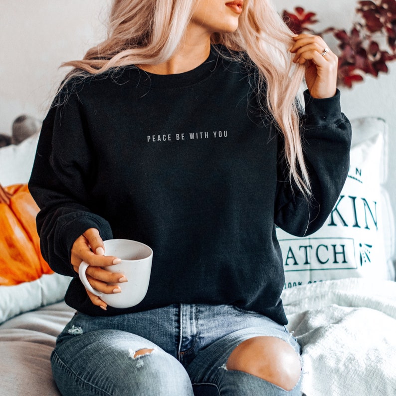 Pu&ograve; includere: Una felpa nera con la scritta "Peace be with you" in lettere bianche. La modella indossa la felpa con jeans blu.