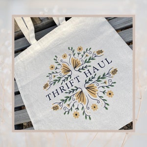 Pu&ograve; includere: Una borsa tote in tela bianca con un design floreale e il testo "THRIFT HAUL" in blu navy.