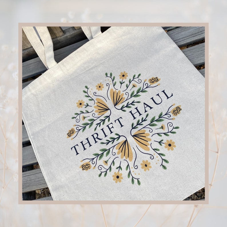 Pu&ograve; includere: Una borsa tote in tela bianca con un design floreale e il testo "THRIFT HAUL" in blu navy.