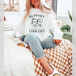 Puede incluir: Sudadera blanca con un gr&aacute;fico de dos libros de dibujos animados tomados de la mano y el texto "Support Your Local Library".