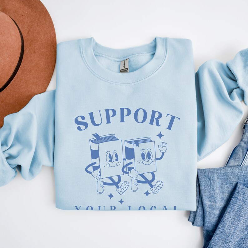 Puede incluir: Una sudadera azul claro con el texto "SUPPORT YOUR LOCAL" en letras azules. Dos personajes de libros de dibujos animados caminan uno al lado del otro, tomados de la mano.