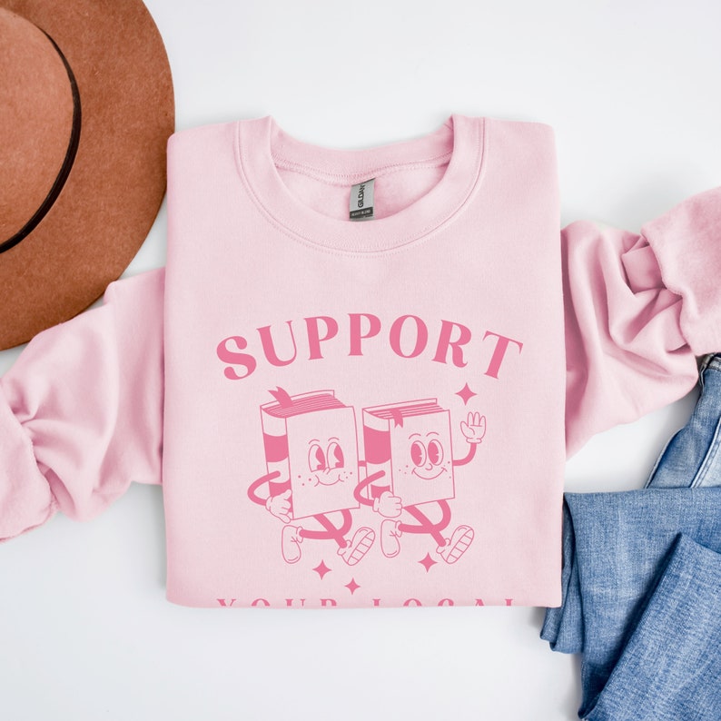 Puede incluir: Sudadera rosa con el texto "SUPPORT YOUR LOCAL" en letras rosas. Dos libros de dibujos animados con caras caminan uno al lado del otro.