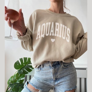 Puede incluir: Una sudadera beige con la palabra "AQUARIUS" impresa en blanco con un peque&ntilde;o coraz&oacute;n debajo.
