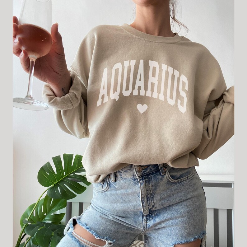 Puede incluir: Una sudadera beige con la palabra "AQUARIUS" impresa en blanco con un peque&ntilde;o coraz&oacute;n debajo.