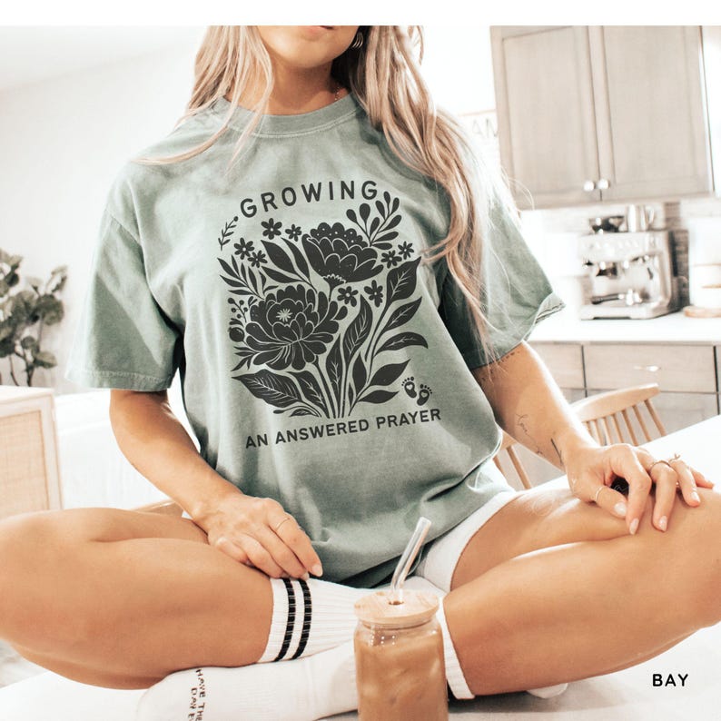 May include: A sage green short-sleeve t-shirt featuring a black graphic of a floral bouquet with the text "GROWING" above the flowers and "AN ANSWERED PRAYER" below, accompanied by a small icon of two baby footprints near the bottom right.