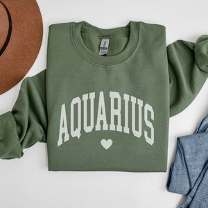 Puede incluir: Una sudadera verde con la palabra "AQUARIUS" en letras blancas en forma de arco con un peque&ntilde;o coraz&oacute;n blanco debajo.