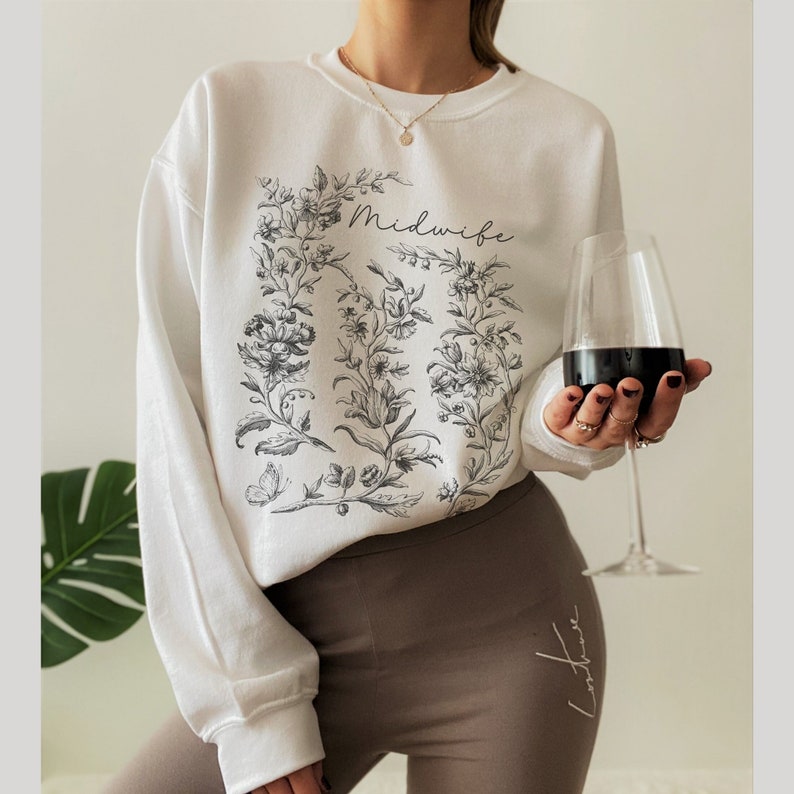 May include: White sweatshirt with a floral design and the word "Midwife" printed on the front. The sweatshirt is being worn by a person with a glass of red wine in their hand.