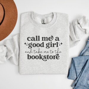 Może przedstawiać: Jasnoszara bluza z okrągłym dekoltem i ciemnoszarym napisem, call me a good girl and take me to the bookstore, ozdobiona małymi czteroramiennymi gwiazdkami. Odzież jest prezentowana obok brązowego filcowego kapelusza i niebieskich dżins&oacute;w.
