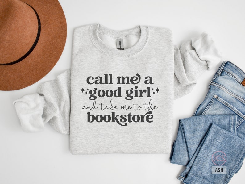 Może przedstawiać: Jasnoszara bluza z okrągłym dekoltem i ciemnoszarym napisem, call me a good girl and take me to the bookstore, ozdobiona małymi czteroramiennymi gwiazdkami. Odzież jest prezentowana obok brązowego filcowego kapelusza i niebieskich dżins&oacute;w.