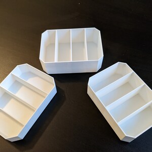 May include: Three white plastic storage containers, each with three compartments. The containers are empty and have a rectangular shape.