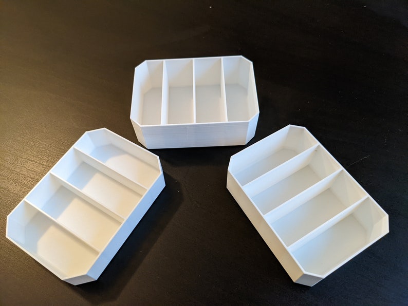 May include: Three white plastic storage containers, each with three compartments. The containers are empty and have a rectangular shape.