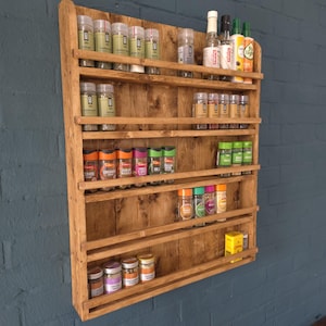 May include: A wall-mounted wooden spice rack with five horizontal shelves. The rack is constructed from light brown stained wood planks and holds various clear plastic spice jars with colourful labels and lids arranged across the shelves.