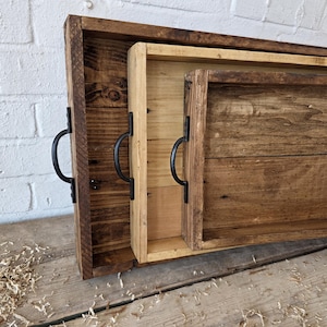 May include: Three rustic wooden serving trays in varying sizes. The trays are rectangular with dark, light, and natural wood tones. Each has a black metal handle. The trays are nested together.