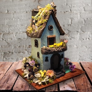 May include: A light blue miniature house sculpture with a dark brown roof covered in green moss. The structure features a small balcony with yellow flowers, a dark arched doorway, and several windows. The base is surrounded by purple and white flowers.