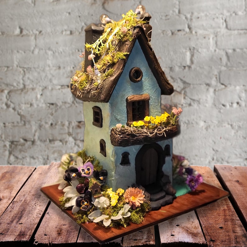 May include: A light blue miniature house sculpture with a dark brown roof covered in green moss. The structure features a small balcony with yellow flowers, a dark arched doorway, and several windows. The base is surrounded by purple and white flowers.