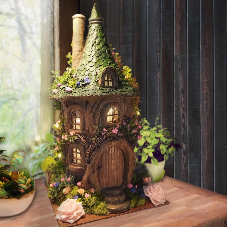May include: A decorative fairy house sculpture featuring a brown tree-trunk base, a textured green shingled roof, and a tall chimney. The structure includes arched windows with warm interior lighting, a wooden door, and artificial moss and pink roses.