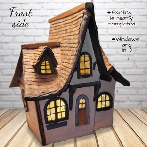 May include: A miniature house sculpture with a brown shingled roof, light gray walls, and dark trim. The structure features illuminated yellow windows and a brown door. Text on the image reads: Front side, Painting is nearly completed, Windows are in :)
