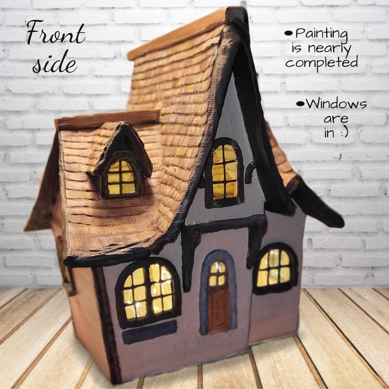 May include: A miniature house sculpture with a brown shingled roof, light gray walls, and dark trim. The structure features illuminated yellow windows and a brown door. Text on the image reads: Front side, Painting is nearly completed, Windows are in :)