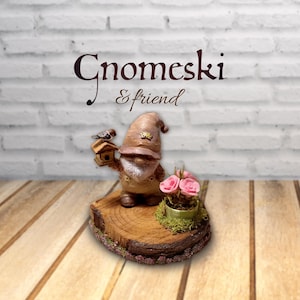 May include: A brown gnome figurine holding a small birdhouse, standing on a circular wood slice base next to a miniature pot of three pink roses. The text Gnomeski & friend is displayed above the figurine in a dark brown script font.